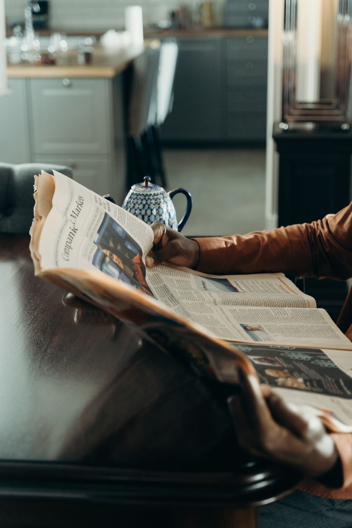 An adult reading a newspaper while sitting at a wooden table indoors, with a teapot in the background.